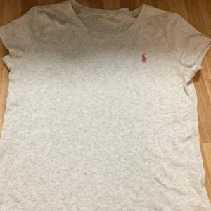 Grå t-shirt från Ralph Lauren - En enkel och stilren grå t-shirt från Ralph Lauren med en liten rosa logotyp på bröstet. T-shirten har korta ärmar och en rund halsringning. köpt från Ohajo