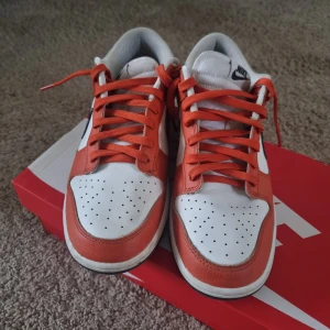 Nike Dunk sneakers i vitt och orange - Ett par Nike Dunk sneakers i vitt och orange med svarta detaljer. Skorna har en vit bas med orange överlägg och svarta swoosh-logotyper med stjärnmönster. De har orange snörning och en vit mellansula med en svart yttersula.