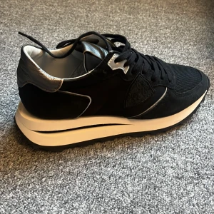 Philippe Model - Ett par svarta sneakers från Philippe Model med vita sulor och svarta snören. Skorna har en silverdetalj på hälen och tungan. De är tillverkade i en kombination av material som ger en stilren och modern look. modell: Tropez haute low Woman. använda 3ggr, nyskick 