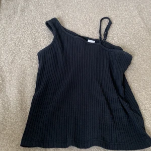 Svart ribbad oneshoulder topp från Lindex - En svart ribbad oneshoulder topp från Lindex. Toppen har en enkel axelbandslös design med en justerbar rem på ena sidan. Tillverkad av ett bekvämt och stretchigt material.