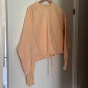 Beige tröja med dragsko - En persikofärgad långärmad tröja med dragsko i nederkanten. Tröjan har ribbade muddar vid ärmslut och en rund halsringning.