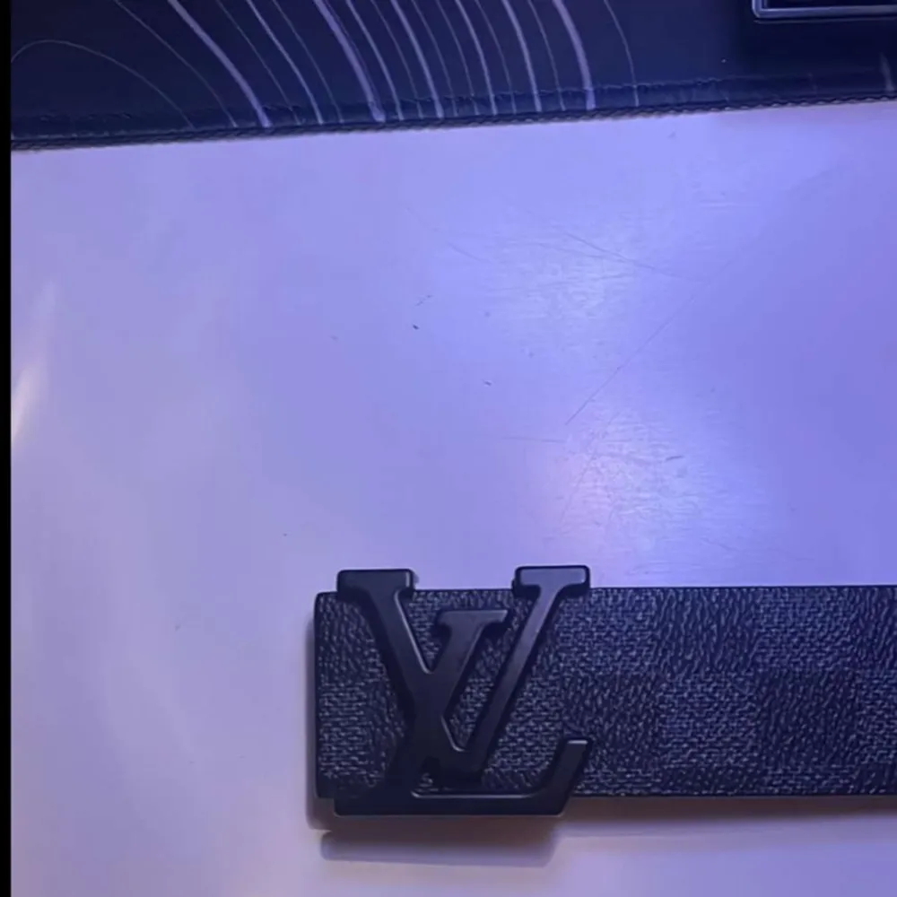 Ett elegant svart bälte från Louis Vuitton med ett klassiskt rutmönster och en svart spänne med LV-logotypen. Bältet är tillverkat av skinn och har en stilren design som passar både till vardags och fest.. Övrigt.