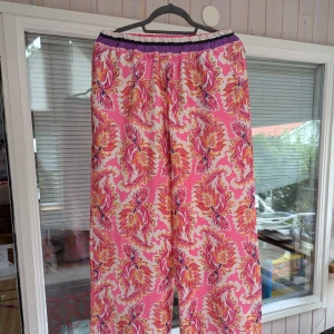 Blommiga byxor från Italien - Ett par färgglada byxor med blommönster i rosa, orange och lila. Byxorna är tillverkade i Italien av 100% polyester och har en elastisk midja för bekväm passform. De har även dekorativa ränder i lila och vitt vid byxbenens slut.