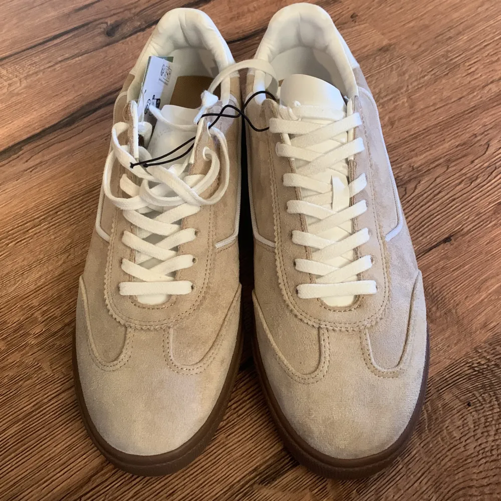 Ett par helt nya beige sneakers från H&M i mocka med vita snören och en brun sula. Skorna har en klassisk design med en bekväm passform. Säljer då de inte passade. Skor.