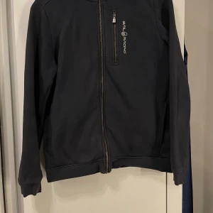 Svart jacka från Sail Racing - Säljer en blå kofta från Sail Racing. Jackan har en dragkedja framtill och en bröstficka med dragkedja. Den är långärmad och har en normal passform. Perfekt för vår och höst!                             Den är köpt från kidsbrandstore för cirka ett år sedan och är i storlek 170