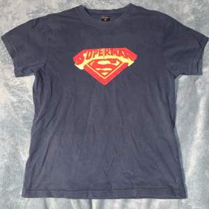 Säljer en cool svart Superman t-shirt med den klassiska Superman-loggan i rött och gult på bröstet. T-shirten är i bra skick och perfekt för alla Superman-fans. Den har korta ärmar och är gjord av bomull, vilket gör den bekväm att bära. Passar perfekt till vardags eller för en avslappnad look.