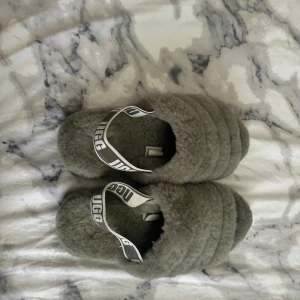 Gråa fluffiga tofflor från UGG - Säljer ett par supermysiga gråa tofflor från UGG. De är fluffiga och har en elastisk rem med UGG-loggan för extra stöd. Perfekta för att hålla fötterna varma och bekväma hemma. Sulan är slitstark och har ett mönster för bättre grepp.