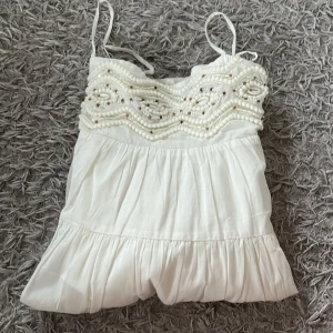 Vit linnetopp med pärldetaljer - En vit klänning med smala axelband och pärldetaljer på överdelen. Klänning är tillverkad i ett lätt och luftigt material och passar Xs och s