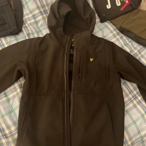 Svart jacka från Lyle & Scott - Säljer en svart jacka från Lyle & Scott i storlek 15/16 år. Jackan har en dragkedja framtill och en liten gul logga på bröstet. Den har en huva och är perfekt för höst- och vårsäsongen. Jackan är i bra skick och har en normal passform. Perfekt för både vardag och lättare utomhusaktiviteter.