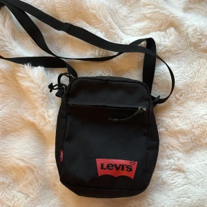 Levis axelväska  - En Levis axelväska i nyskick, en liten ficka på utsidan och en liten med dragkedja. Väskan är i nyskick och funkar jättebra att ha med sig vart än man är. 