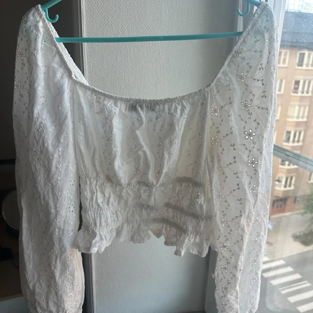 En vit blus från Chiquelle med broderade detaljer och elastisk midja. Blusen har långa ärmar och volangdetaljer vid nederkanten.. Toppar.