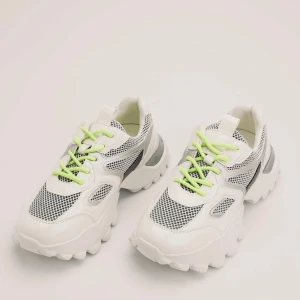 Vita sneakers med gröna snören - Ett par vita sneakers med gröna snören och svarta detaljer. Skorna har en tjock sula och är tillverkade av syntetmaterial med meshpaneler för andningsförmåga.