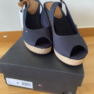 Svarta sandaler från Tommy Hilfiger - Ett par svarta sandaler från Tommy Hilfiger tillverkade av ekologisk bomull. Sandalerna har en kilklack med flätad detalj och en öppen tådesign.