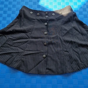 Svart kortkjol med knappar - En svart kortkjol med tunna vita ränder och knappar framtill. Kjolen har en A-linjeform och är tillverkad i ett lätt material.