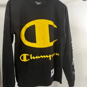 En svart långärmad tröja från Champion i samarbete med Supreme. Tröjan har en stor gul Champion-logga på framsidan och Supreme-tryck på ena ärmen. Den är tillverkad i bomull och har en normal passform.