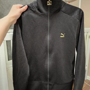 Svart jacka från Puma - En svart jacka från Puma med dragkedja framtill. Jackan har en hög krage och är tillverkad av 66% polyester och 34% bomull. Den har en enkel design med Puma-logotypen i guld på bröstet.