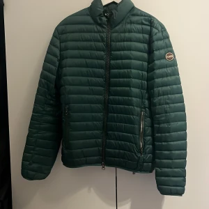 Grön pufferjacka från Colmar - En grön pufferjacka från Colmar med dragkedja framtill och två dragkedjeförsedda fickor. Jackan har en quiltad design och en hög krage för extra värme. Size S