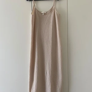 En begie trikå klänning  - En krinklad trikå klänning i färgen beige. Klänningen är vadlång och är ärmlös och har smala reglerbara axelband. Och har en liten ihopsydd slits på baksidan. Har bara testats. 