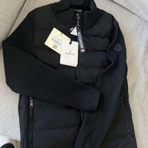 Moncler Cardigan sport black lycra - Denna svarta Moncler-cardigan i sportlycra kombinerar stil och funktion. Med Monclers signaturlogga och en figurnära siluett ger den ett sofistikerat och sportigt utseende. Materialet är stretchigt och andas, vilket gör den bekväm för både vardag och