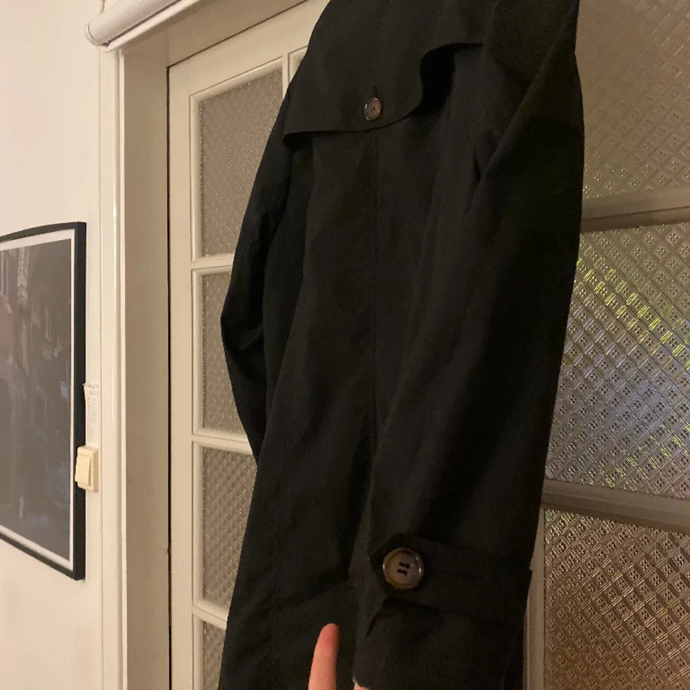 En fin svart kappa/trenchcoat med bruna knappar och coola detaljer. Storleken skulle jag uppskatta till en L. Köpt secondhand, men har inga tecken på användning.. Jackor.