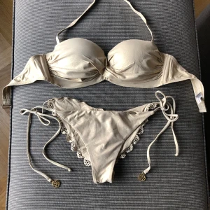 Lulifama Bikini strl Medium i Guld med vackra detaljer! - Underbar bikini från Miamimärket Lulifama i Storlek Medium köpt i deras butik i Miami. Nypris $200, ca 2000kr. Uttagbara push-up inlägg, även Bandeau då störret runt halsen kan tas bort/ på enkelt. Lite spår av användning på metalldetaljer