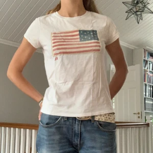 Ralph Lauren Tshirt - Ralph Lauren tshirt i offwhite och med amerikanska flaggan. Storlek M men en liten M enligt mig. Säljer för 150 kr. Gratis frakt vid köp av två eller fler plagg.