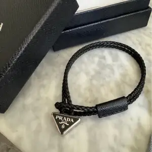 Svart armband med ikoniska Prada triangeln. Armbandet i svar saffiano läder.  Inköpt 2020 på Pradas hemsida. Använd Max 10 gånger. Orginalbox samt originalpåse kan fås med vid önskemål. Kvitto finns också. 