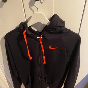 En Nike jacka som är svart med orangea drag och väldigt skön och lite oversized funkar o träna i och styla snyggt 