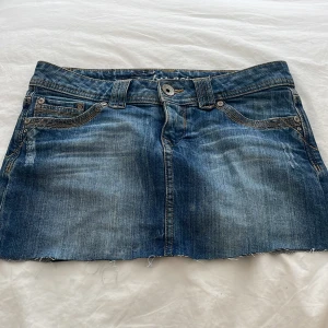 Blå jeanskjol - En blå jeanskjol med slitna detaljer och rå kant. Kjolen har fram- och bakfickor samt bälteshällor. Den är kort och har en knapp- och dragkedjestängning framtill.