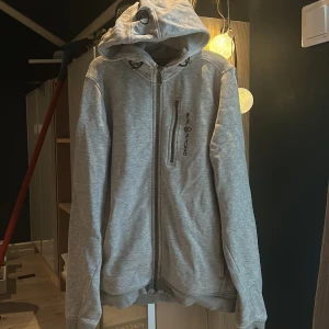 Grå sail racing hoodie - Säljer min gråa sail racing tröja som tyvärr inte kommer till användning längre. Den har ett litet hål på armen som är så litet att de inte märks å även som ett litet sträck som inte syns om man har på blixt annars en fin tröja!skriv för fler bilder