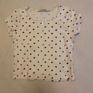 T-shirt med små jordgubbar  - Jättegullig t-shirt med små jordgubbar på. Spets i linningen och en liten rosett. Osliten, använd ca 5 gånger.
