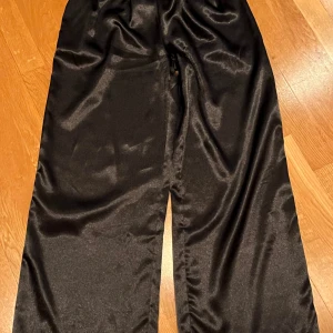 Glansiga svarta vida satin byxor - Såå sköna! Och coola! Blanka svarta vida byxor med resår i midjan. Fina detaljer och fin linning gör dem ”klädda”, resår baktill i midjan. Perfekt längd till en som är ca 168cm lång. Stl S. Nya! Från Isay. Nypris 799kr.