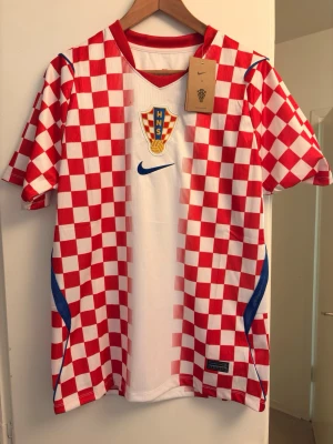 Kroatien fotbollströja Nike röd/vit - Säljer en officiell Kroatien fotbollströja från Nike med klassiskt rödvitt schackrutigt mönster, blå detaljer och HNS-emblem på bröstet. Tillverkad i Dri-FIT material som andas, perfekt för träning eller match.. 