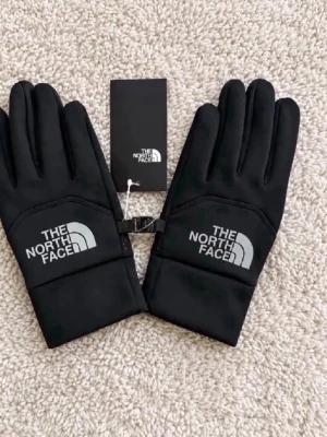 Svarta handskar från The North Face - Snygga svarta handskar från The North Face med logga i vitt på ovansidan. Handskarna har en stilren design och är tillverkade i ett mjukt syntetmaterial som passar perfekt för kyliga dagar. De har en smidig passform och kan enkelt kopplas ihop med ett spänne.