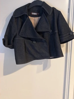 Kortärmad jeansjacka från DENIM CO - Mörkblå kortärmad jeansjacka från DENIM CO med bred krage, dubbelknäppning och stora knappar. Jackan har en boxig, croppad passform och dekorativa detaljer på ärmarna. Perfekt för en trendig och avslappnad look.
