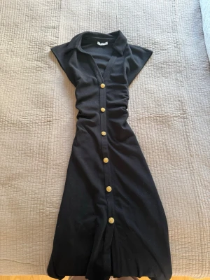 Svart långklänning med guldknappar - Stilren svart långklänning med krage och dekorativa guldknappar längs hela framsidan. Klänningen har v-ringning och är ärmlös, vilket ger en elegant och tidlös look. Perfekt för dig som gillar klassiska detaljer och enkel design.