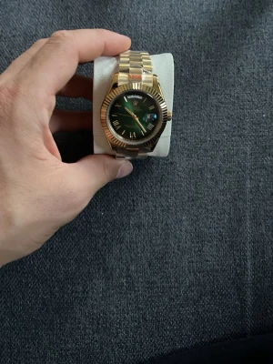 Guldig klocka med grön urtavla - Stilren klocka med guldig länk och grön urtavla. Klockan har romerska siffror och datumvisning, samt en markerad räfflad kant runt urtavlan. Perfekt accessoar för dig som vill sticka ut med en lyxig känsla.