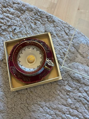 Lyxig tekopp med fat i guld och vinrött - Säljer en elegant tekopp med matchande fat i porslin. Koppen och fatet har vinröd bas med detaljerade guldfärgade mönster och insida i vitt med gulddetaljer. Handtaget har en unik dekoration i silverfärg. Perfekt för dig som gillar klassisk och lyxig design.
