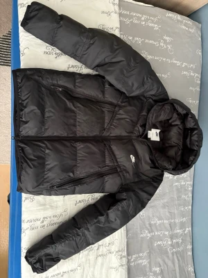 Svart pufferjacka från Nike med huva - Säljer en svart pufferjacka från Nike med huva och två stora dragkedjeförsedda fickor framtill. Jackan har en klassisk pufferdesign med breda kanaler och Nike-logga på bröstet. Perfekt för kalla vinterdagar och riktigt snygg streetstil.