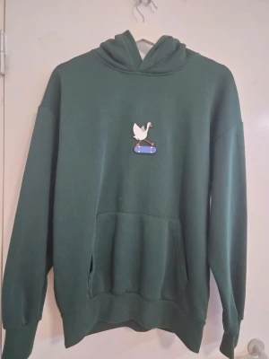 Mörkgrön hoodie med gås-tryck - Mörkgrön hoodie med huva och känguruficka framtill. På bröstet finns ett tryck av en vit gås som åker skateboard. Hoodien har ribbade muddar vid ärmar och nederkant. Perfekt för dig som gillar streetwear och vill sticka ut med ett unikt motiv.