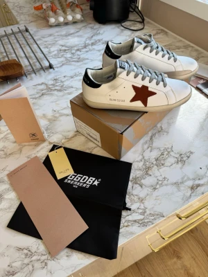 Golden goose.  - Snygga vita sneakers från Golden Goose med grå skosnören, svart häl och en brun stjärna på sidan. Klassisk låg modell med GGDB-detaljer och robust sula. Perfekta för dig som vill ha en trendig och unik look. Stl 39 = 25 cn men tänk på att dom är smala i storleken. Alla tillbehör medföljer. 