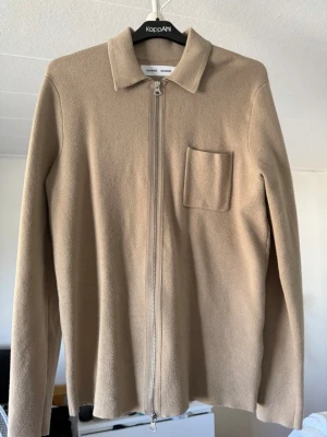 Beige overshirt från Samsøe Samsøe - Stilren beige overshirt från Samsøe Samsøe med rak passform, klassisk krage och dragkedja framtill. En bröstficka på vänster sida ger en clean look. Tillverkad i mjukt material som känns skönt mot huden. Perfekt att slänga över en t-shirt.
