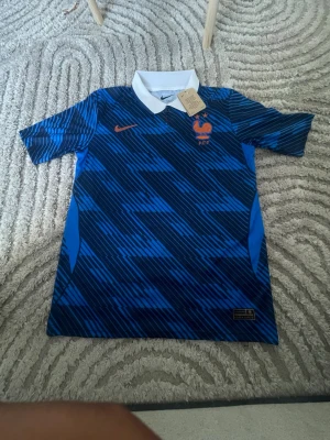 Blå Nike fotbollströja Frankrike L - Säljer en blå fotbollströja från Nike med Frankrikes landslagsmärke och orange detaljer. Tröjan har ett grafiskt mönster i olika blå nyanser, vit krage och är i slim fit-modell. Perfekt för match eller träning!