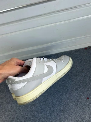 Nike vita och gråa sneakers - Snygga Nike sneakers i vitt och ljusgrått med klassisk swoosh på sidan. Skorna har lågt skaft, vita skosnören och en beige/vit sula. Tillverkade i läder och textil för en clean och stilren look som passar till det mesta.