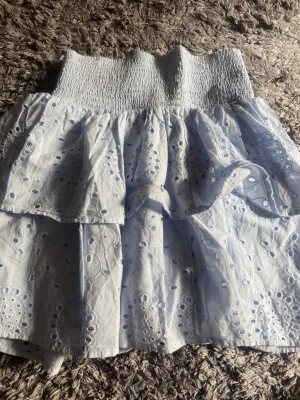 Ljusblå volangkjol med broderi - Supersöt ljusblå kortkjol med volanger och broderade detaljer. Kjolen har en smockad midja som ger en bekväm passform och ett romantiskt uttryck. Perfekt för vår och sommar när du vill ha en luftig och trendig look. 170 i storlek och dessutom ny och oanvänd. 