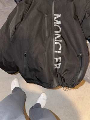 Svart vindjacka från Moncler - Snygg svart vindjacka från Moncler med stor logga i vitt längs dragkedjan. Jackan har flera dragkedjeförsedda fickor och justerbar nederkant. Materialet är tunt och vindtätt, perfekt för blåsiga dagar. Klassisk Moncler-detalj på insidan.