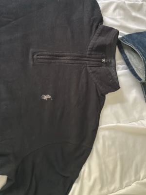 Svart halvzip från Polo Ralph Lauren - Svart långärmad tröja från Polo Ralph Lauren med halv dragkedja och klassisk broderad logga på bröstet. Tröjan har hög krage och är tillverkad i mjukt material som känns skönt mot huden. Perfekt för en stilren och avslappnad look.