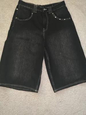 Svarta denimshorts med kontrastsömmar - Säljer ett par svarta denimshorts med vita kontrastsömmar och klassisk femficksdesign. Shortsen har normal passform och raka ben, perfekta för en avslappnad stil. Materialet är slitstarkt och passar bra till sommaren.