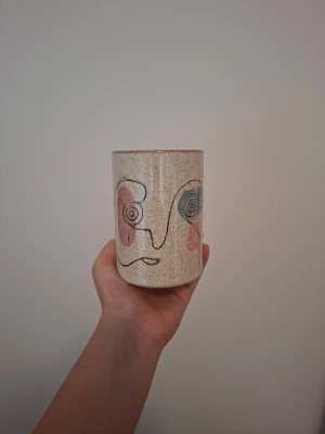 Keramikkopp med abstrakt ansikte - Unik keramikkopp i beige med handmålat abstrakt ansikte i svart, rosa och blått. Koppen har en rak form och konstnärlig design som sticker ut. Perfekt som dekoration i hemmet.