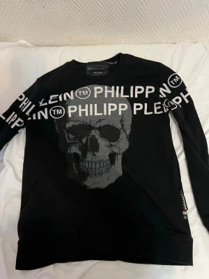 Svart sweatshirt från Philipp Plein - Cool svart sweatshirt från Philipp Plein med stort glittrigt dödskalletryck och vit logga över bröstet och ärmarna. Tröjan har rund halsringning och långa ärmar. Perfekt för dig som gillar streetwear och statementplagg.